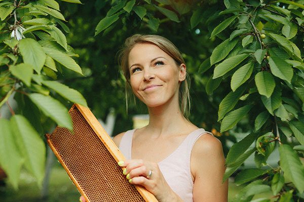 Young woman outdoors standing amongst some low branches, smiling and holding a frame of a beehive - source of many of the ingredients used in Unbeelievable Health's natural supplements